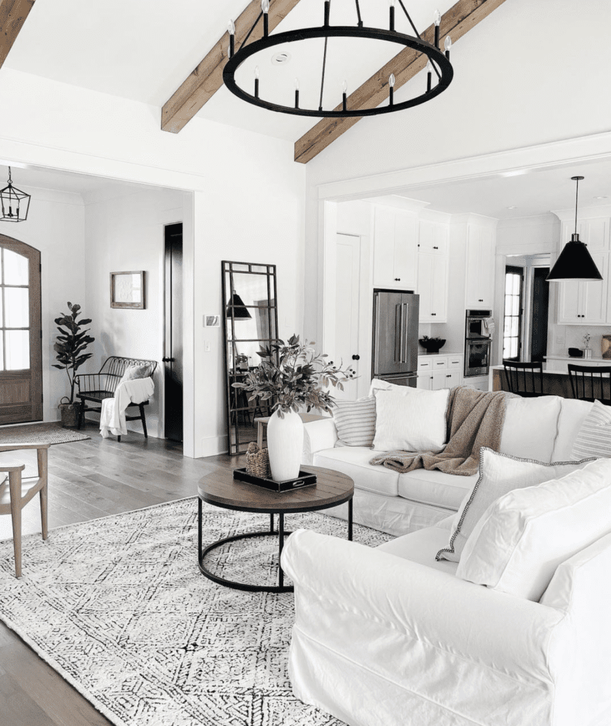 An all-white modern farmhouse living room. The accent pieces are the black wagon wheel metal chandeliers and the dark round wood coffee table