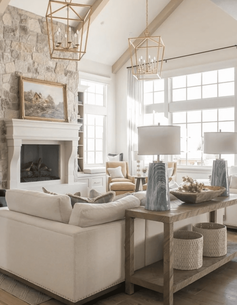 A modern farmhouse living roon in pastel colors. There are two gold lantern chandeliers and a vintage frame with a landscape painting in front of the cozy fireplace