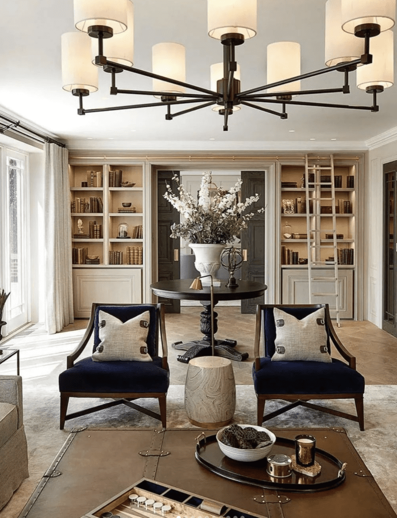 A modern elegant farmhouse living room. There are two blue velved chairs with a table and flowers behind them. Hanging from the ceiling a black metal heavy duty chandelier