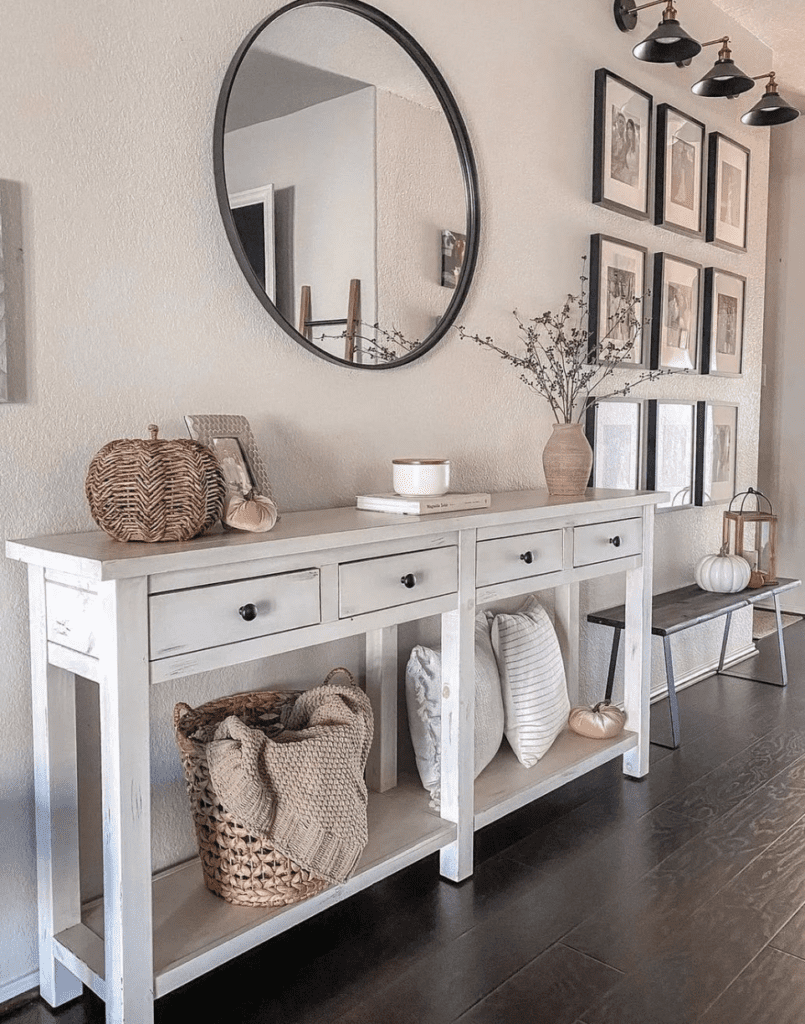 A modern farmhouse console table in distressed white wood. There is a round mirror hanging on the wall.