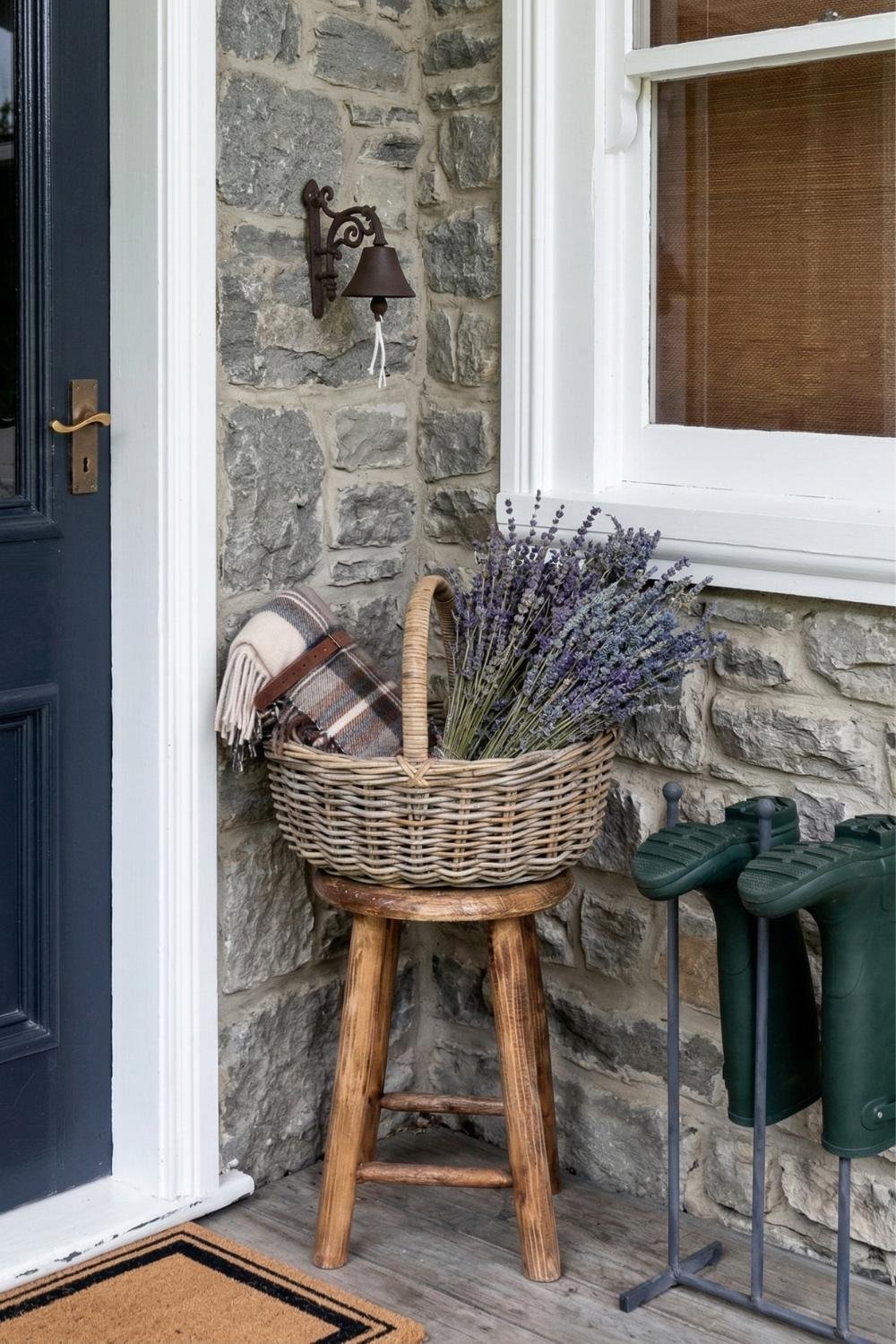 Rustic stone cottage porch decor featuring a wicker basket of lavender on a wooden stool and green rubber wellington boots.