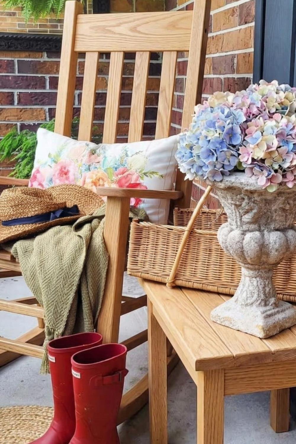 Rustic cottage porch detail with a light wood rocking chair, red rain boots, and a stone urn filled with blue hydrangeas.