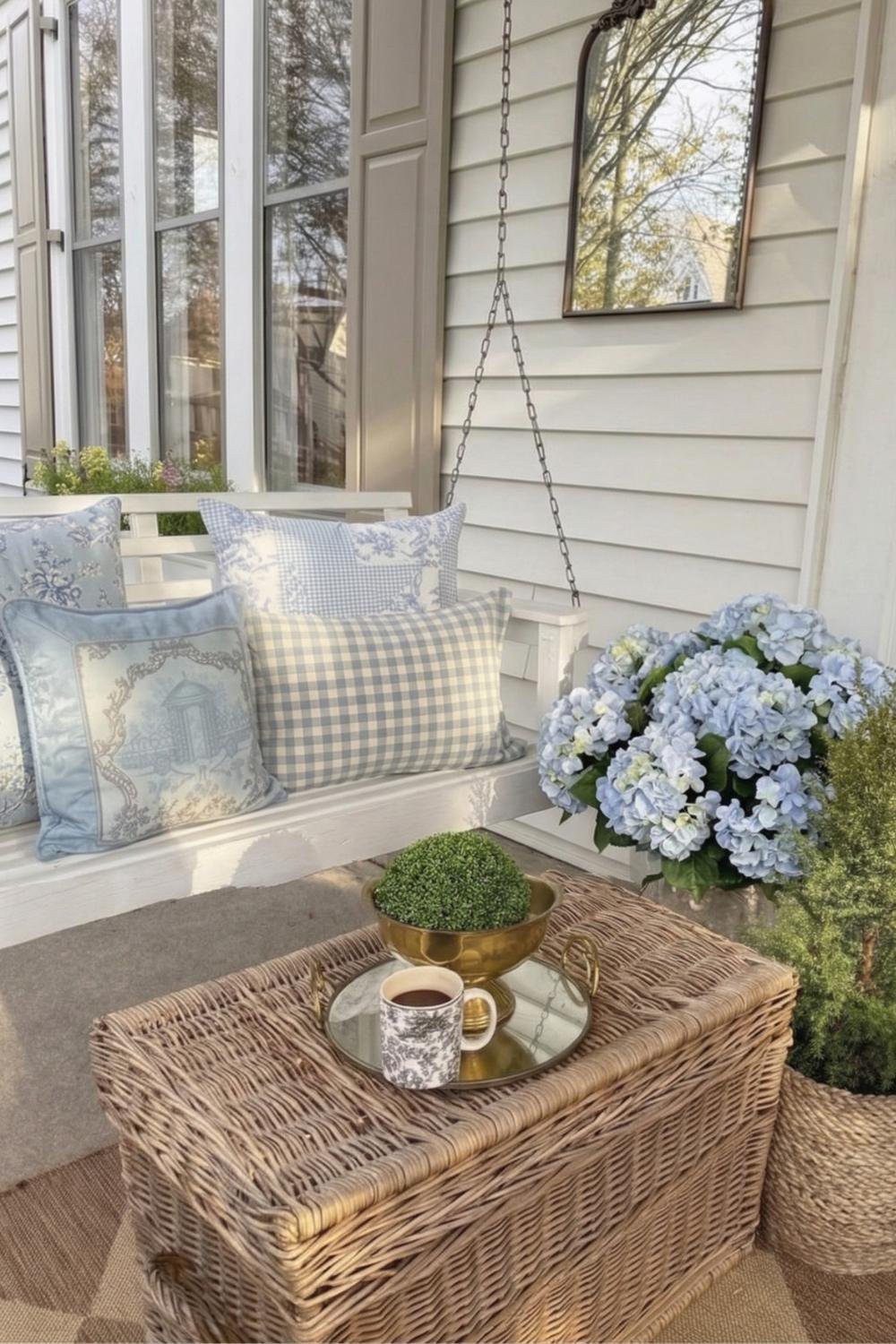 Cozy cottagecore porch swing with light blue vintage patterned pillows, a large hydrangea bush, and a wicker coffee table.