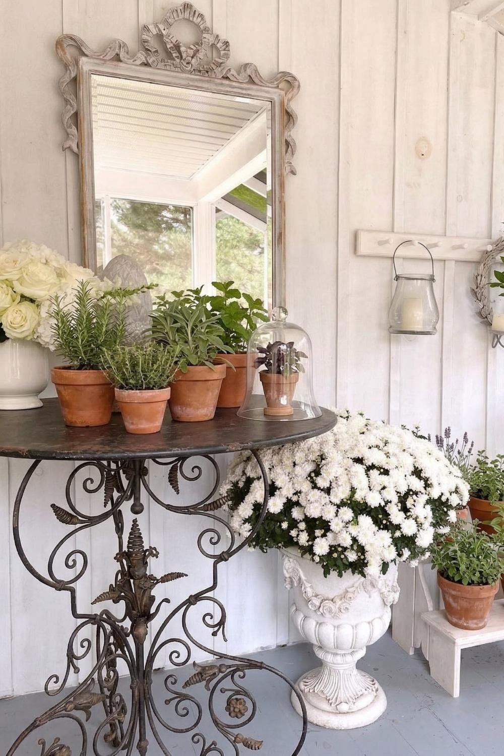 Vintage cottage porch decor with a wrought iron console table, terracotta potted herbs, and a large white floral arrangement in a stone urn.