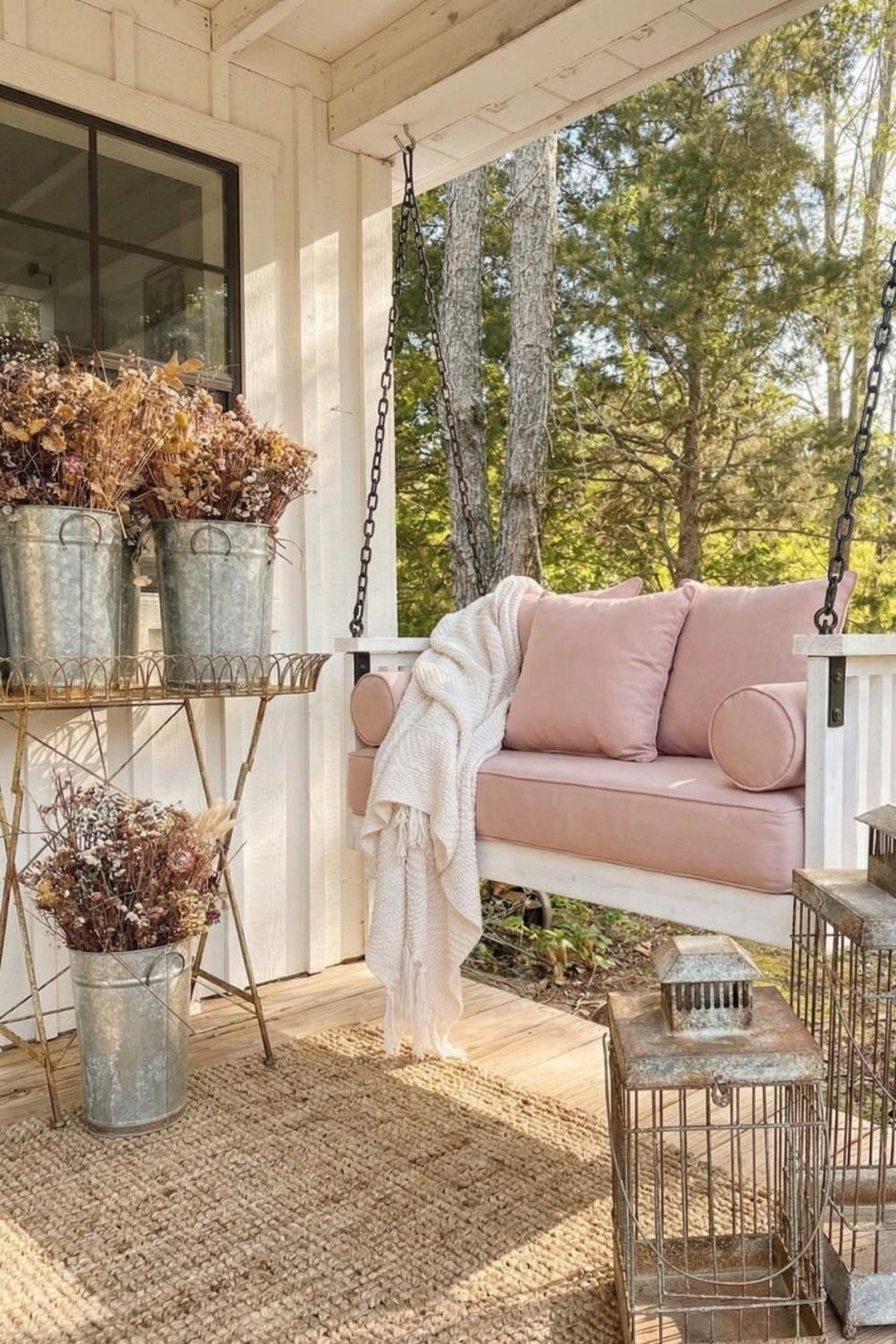 Shabby chic porch swing with dusty pink cushions, a cream throw blanket, and a large jute rug on a sunny wooden deck.