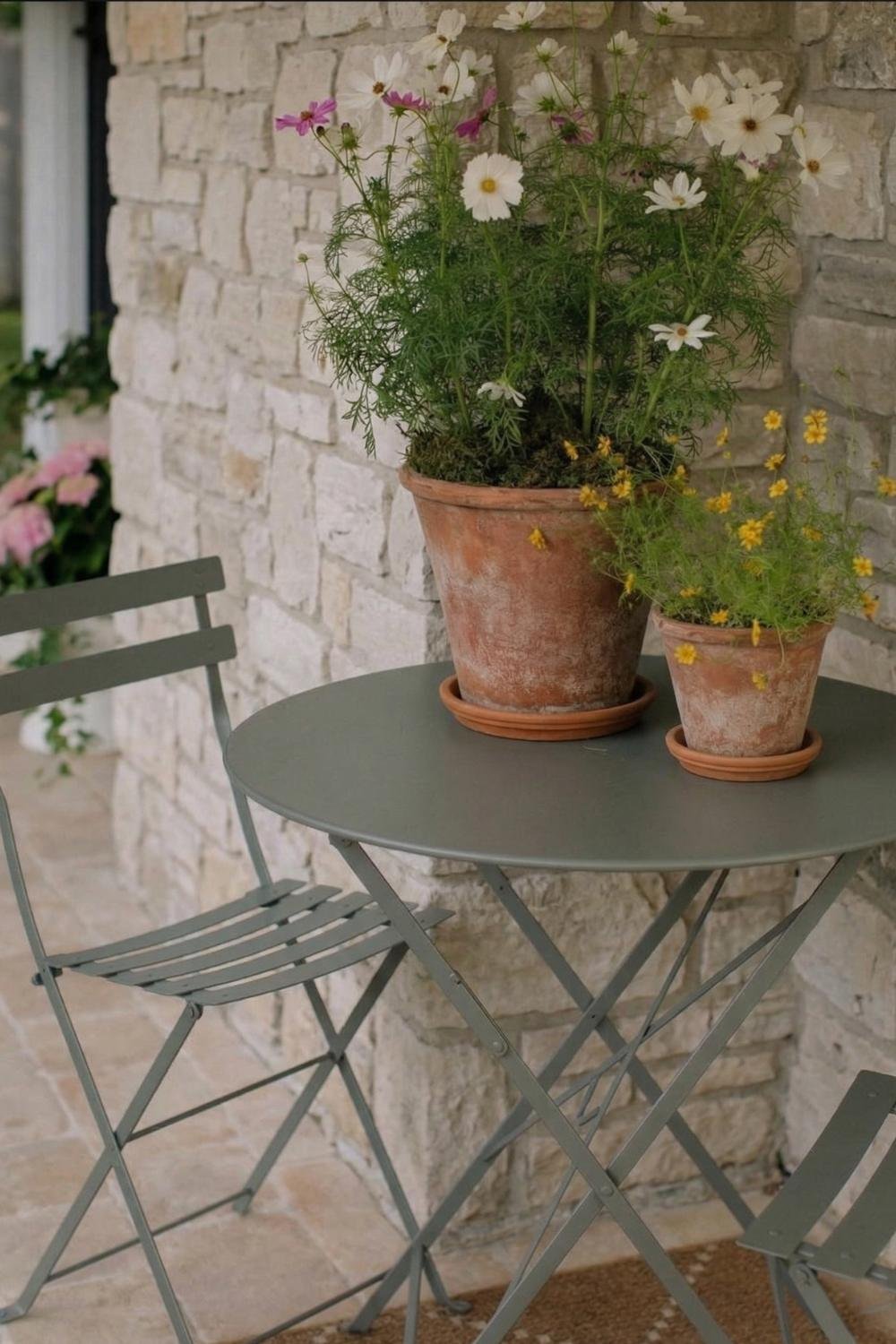 Parisian-style cottage porch bistro set with a grey metal table and chairs against a light stone wall with terracotta wildflowers.